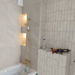 Kompleksowe usługi glazurnicze - Wykończona łazienka z beżowymi płytkami, złotą armaturą i podświetlanymi niszami. Widoczne narzędzia i chemia budowlana. Ukończona obudowa WC z płytkami.