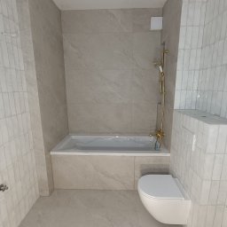 Kompleksowe usługi glazurnicze - Jasna łazienka po remoncie: beżowe płytki imitujące kamień na ścianach i podłodze, biała wanna z obudową, toaleta podwieszana i złota armatura.