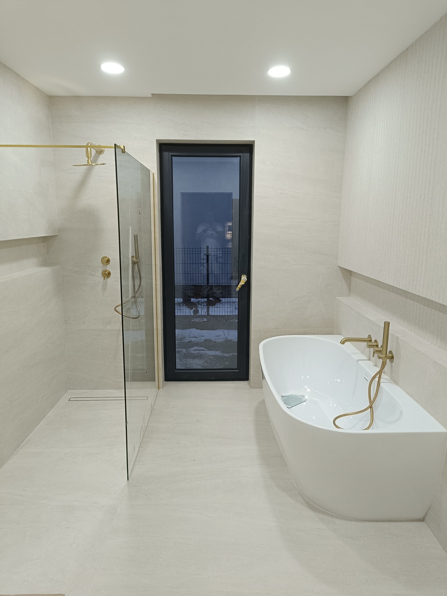 Nowoczesna łazienka z kabiną prysznicową typu walk-in, wolnostojącą wanną i złotą armaturą. Jasne płytki na ścianach i podłodze tworzą spójną, minimalistyczną przestrzeń.