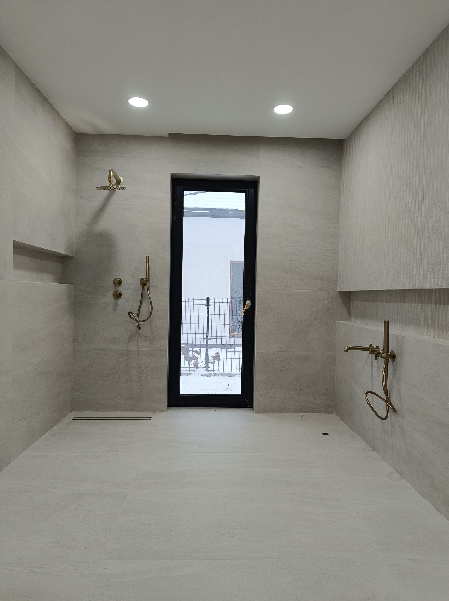 Wykończona łazienka w jasnych odcieniach szarości z złotą armaturą. Widoczne płytki na ścianach i podłodze, kabina prysznicowa typu walk-in oraz okno z czarną ramą.