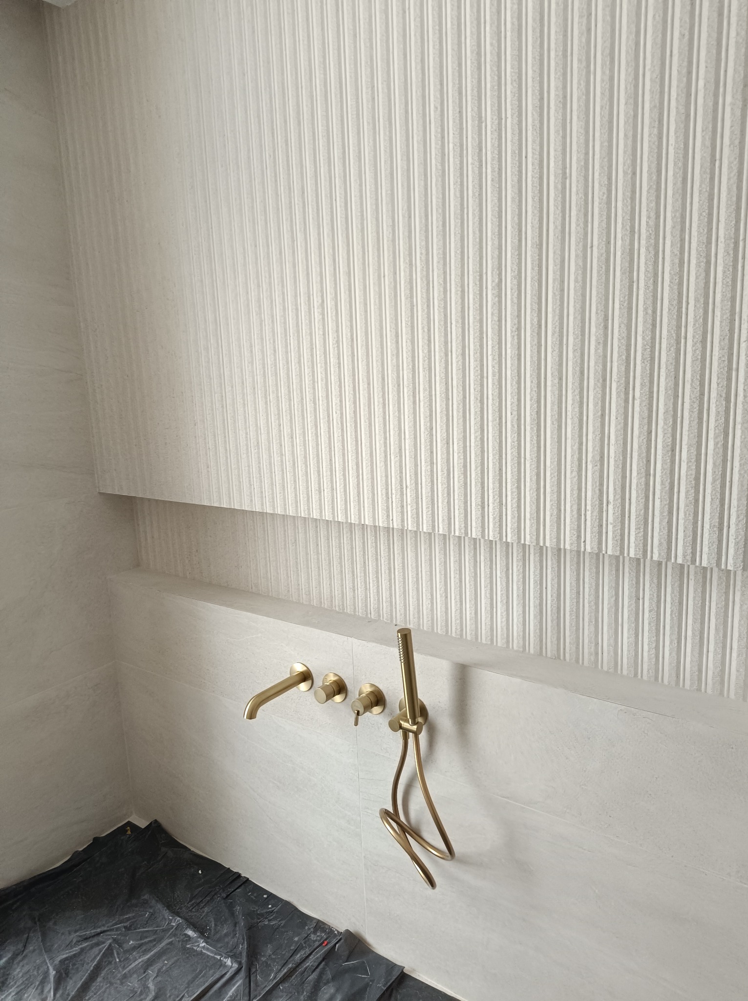 Wykończona ściana w łazience z dekoracyjną, białą glazurą w pionowe prążki i złotą armaturą. Podłoga zabezpieczona folią malarską.