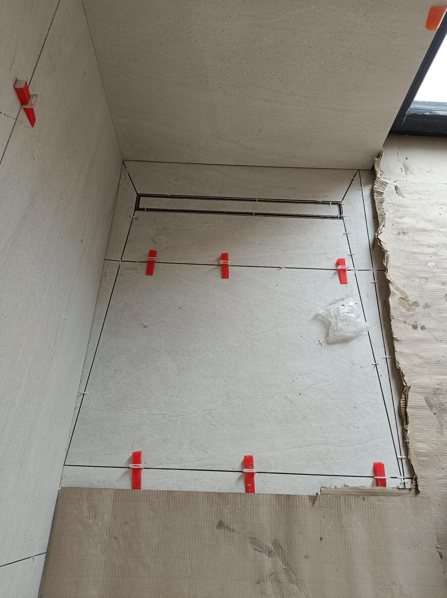 Jasne płytki podłogowe w trakcie układania, z użyciem czerwonych klipsów poziomujących i czarnych listew. Widoczny odpływ liniowy i karton ochronny.