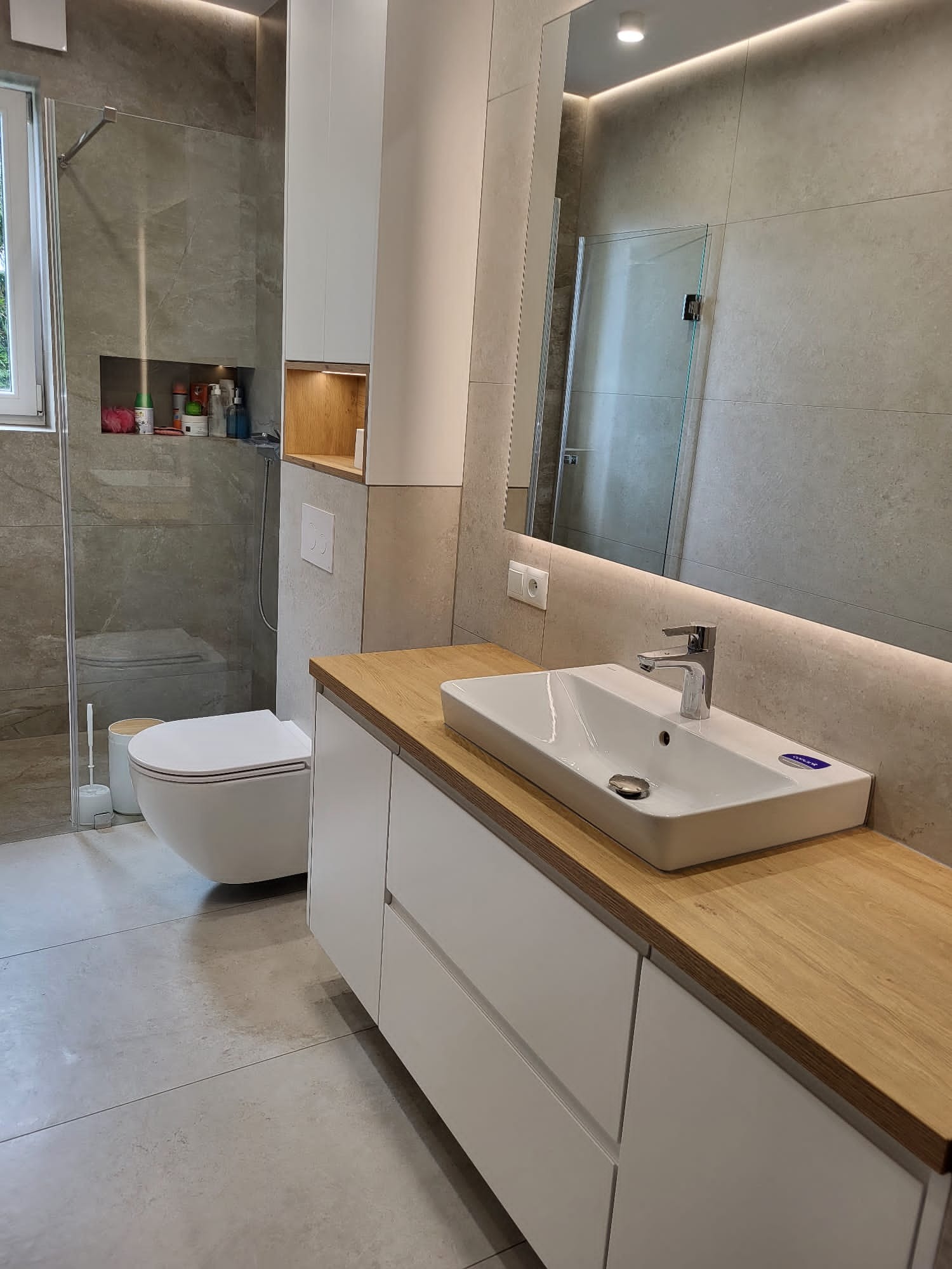 Nowoczesna łazienka z szarymi płytkami, białymi meblami i drewnianym blatem. Umywalka nablatowa, lustro z podświetleniem LED. Minimalistyczny design.