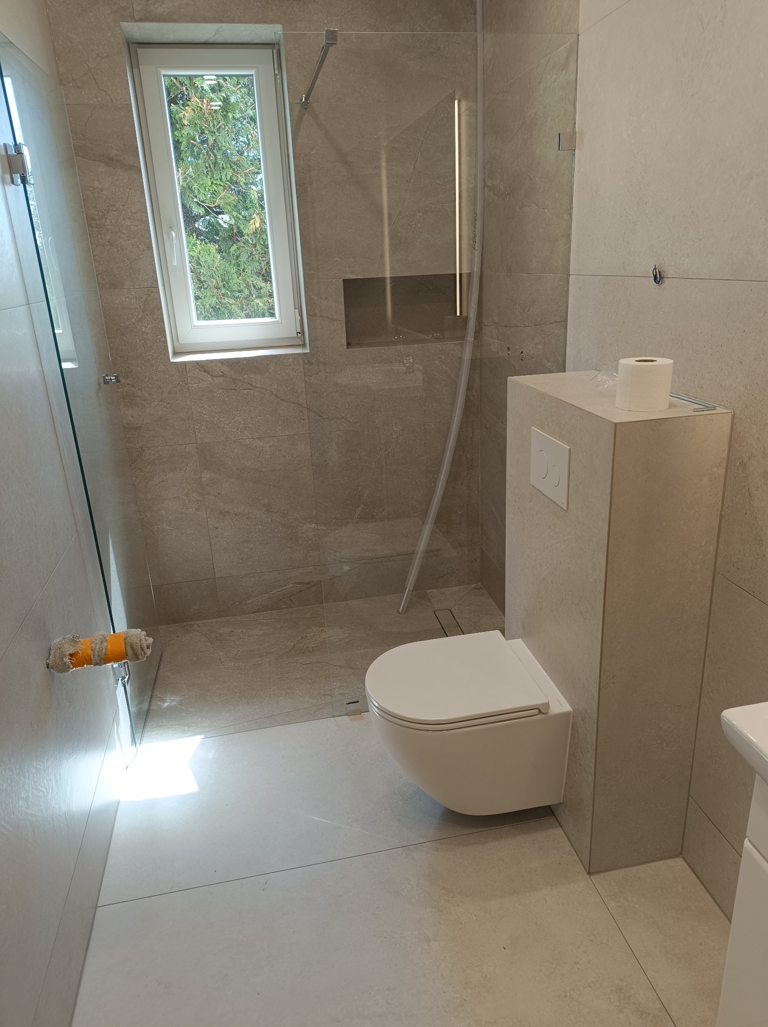 Wykończona łazienka z beżowymi płytkami, wiszącą toaletą i kabiną prysznicową ze szklaną ścianką. Widoczne okno i rolka malarska na ścianie.