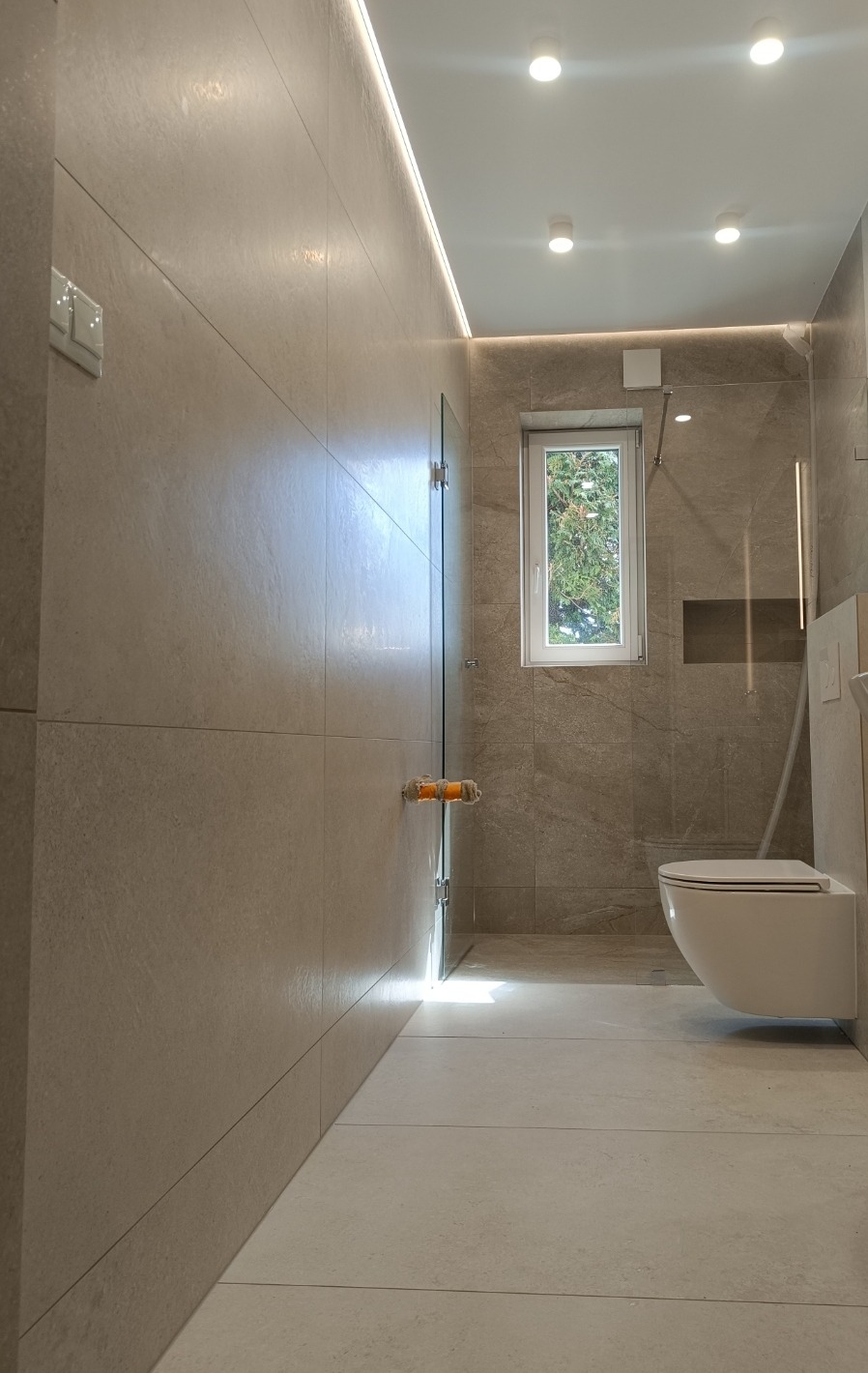 Jasna łazienka z beżowymi płytkami, kabiną prysznicową ze szklanymi drzwiami i podwieszaną toaletą. Minimalistyczny design z oświetleniem LED.