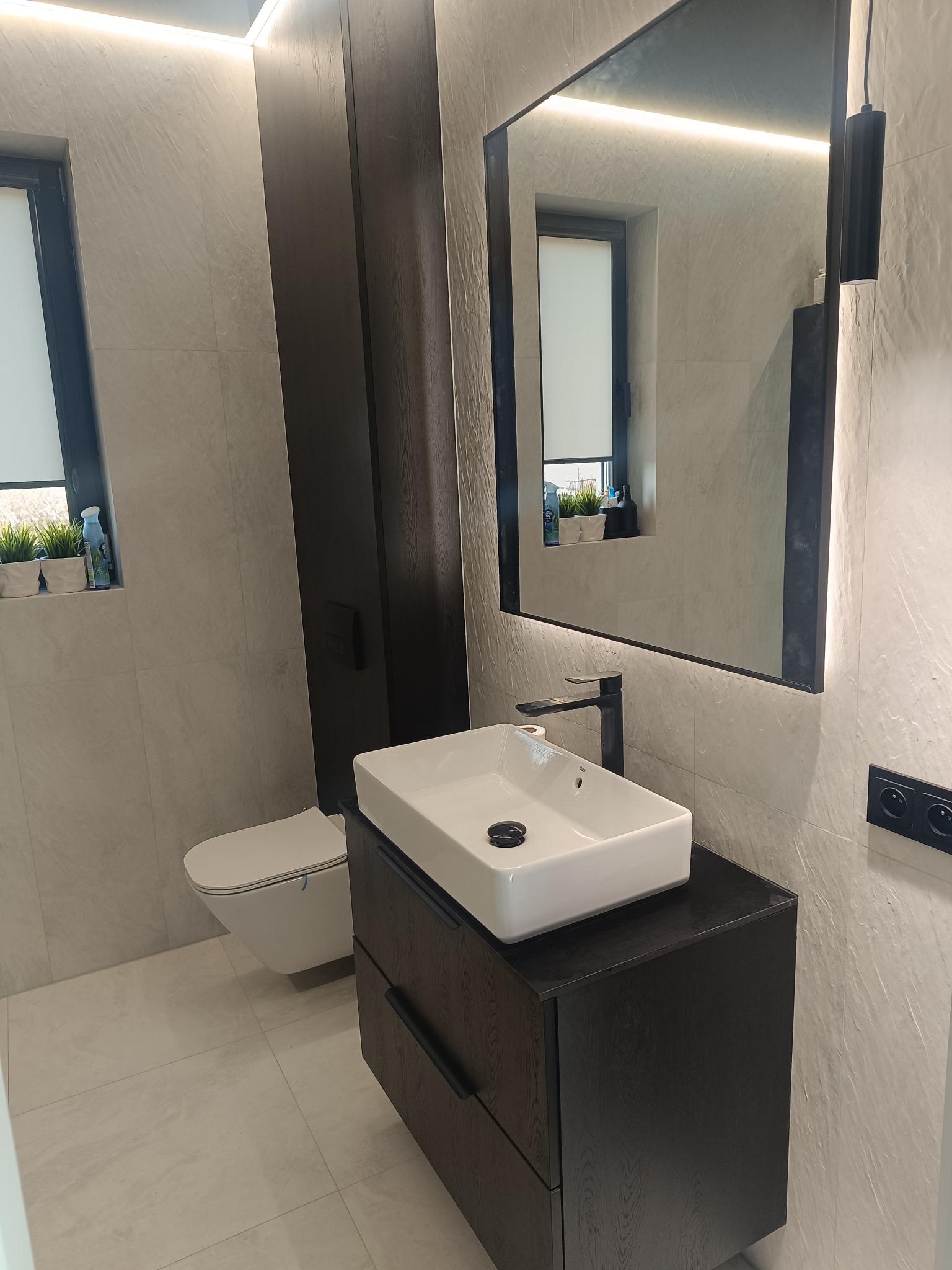 Nowoczesna łazienka z białymi płytkami, wiszącą toaletą i umywalką na szafce z ciemnego drewna. Lustro z podświetleniem LED. Minimalistyczny design.