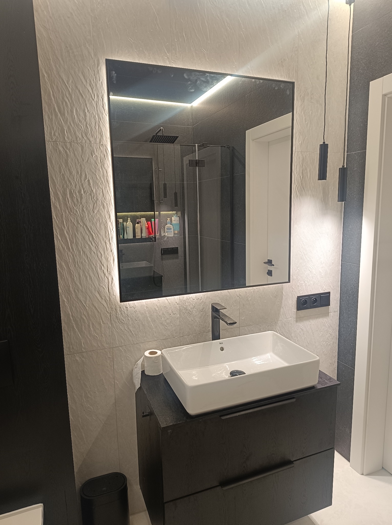 Nowoczesna łazienka z umywalką nablatową, lustrem z podświetleniem LED i płytkami imitującymi kamień. Widoczna kabina prysznicowa w tle.