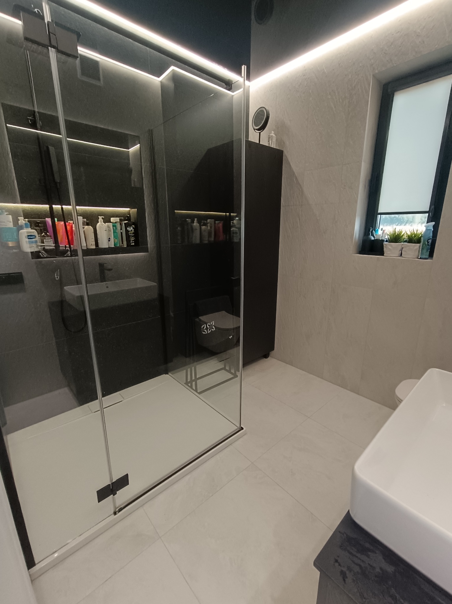 Nowoczesna łazienka z kabiną prysznicową typu walk-in, jasnymi płytkami podłogowymi i ściennymi, oraz oświetleniem LED. Minimalistyczny design z akcentami czerni.