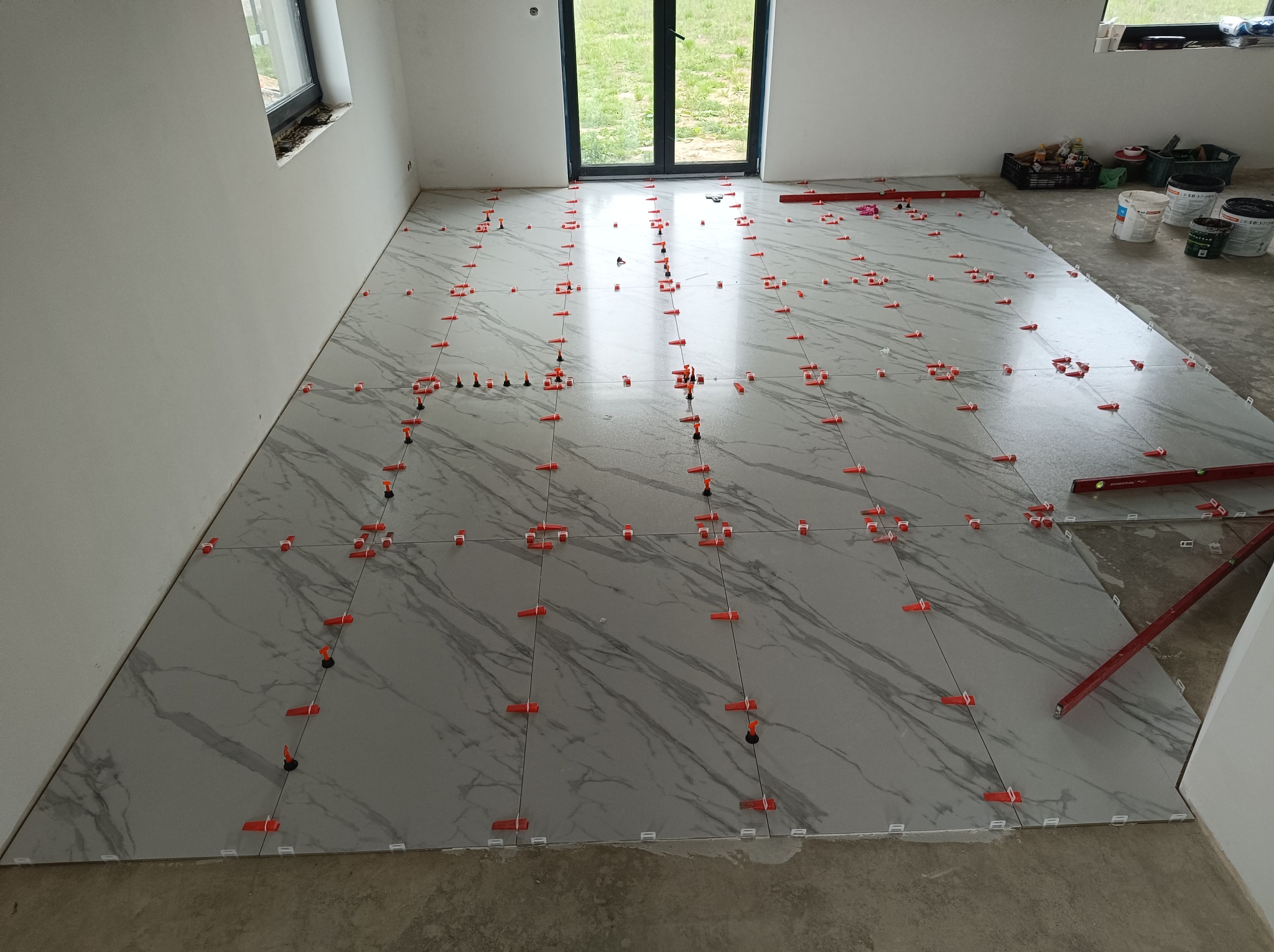 Układanie płytek podłogowych z użyciem systemu poziomowania. Szare płytki imitujące marmur, czerwone klipsy. Częściowo wykończona podłoga w jasnym pomieszczeniu.