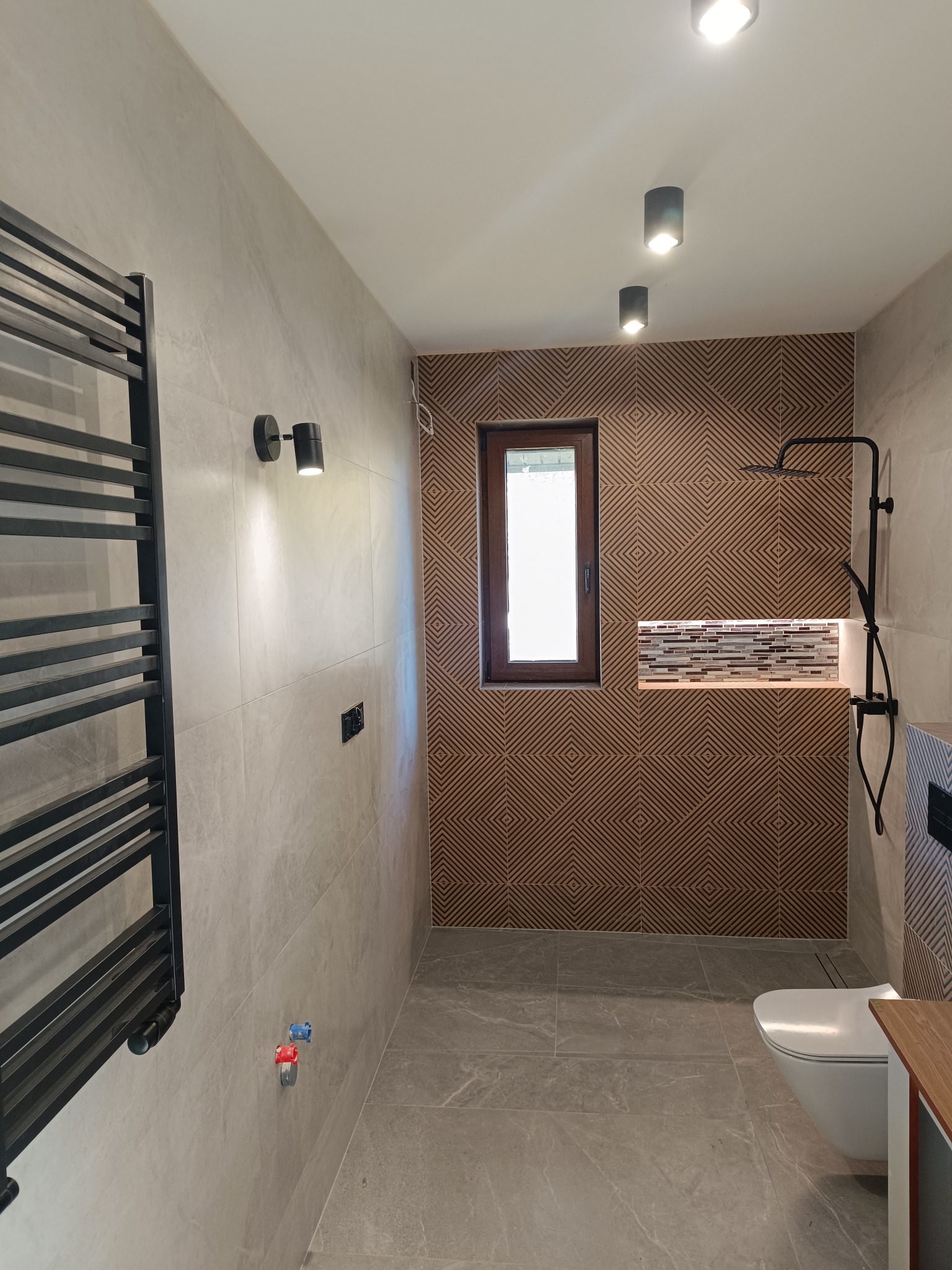 Wykończona łazienka z szarymi płytkami, dekoracyjną ścianą z drewnianymi panelami, czarną armaturą i grzejnikiem drabinkowym. Nowoczesny design.