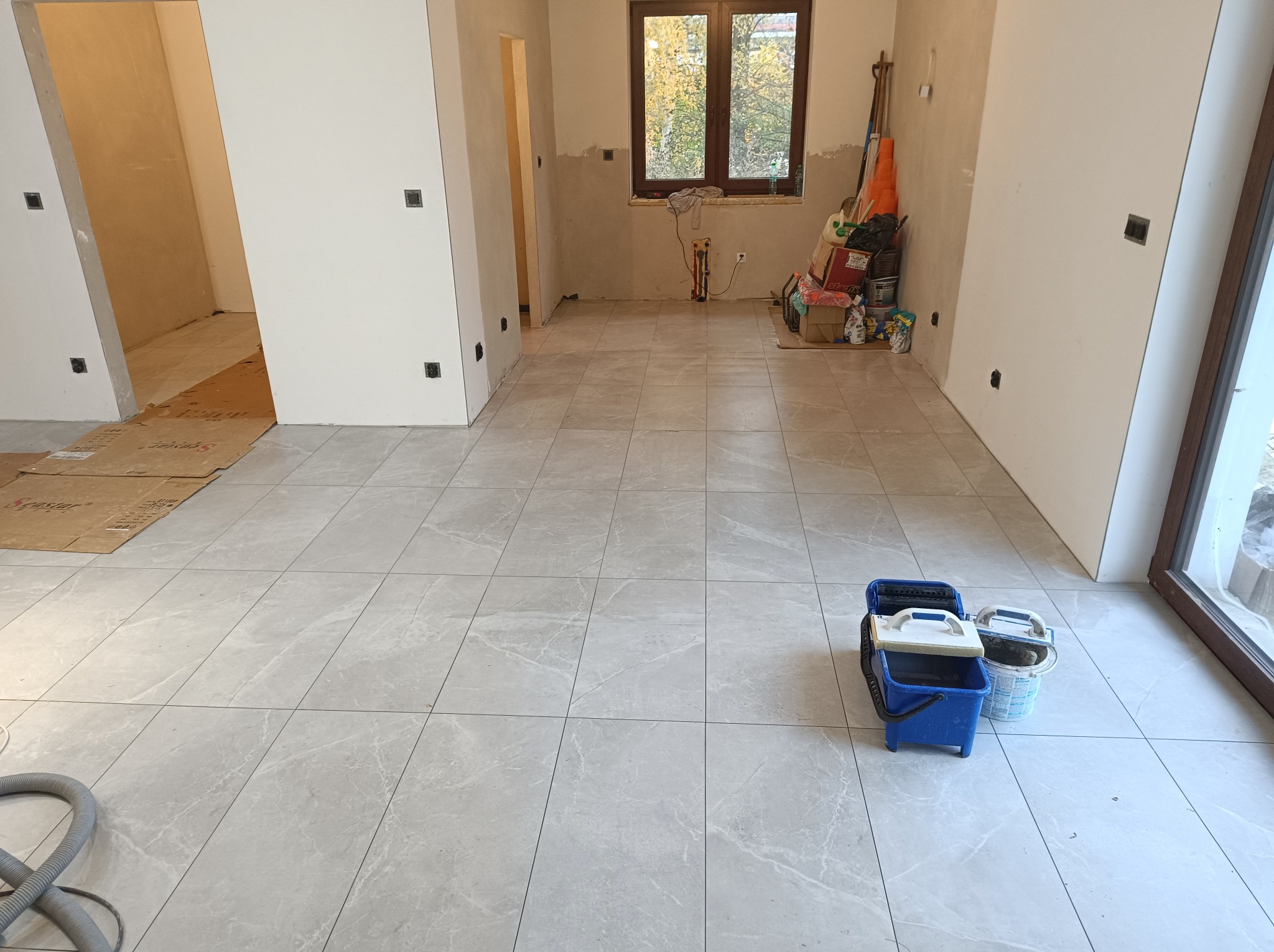 Jasnoszara, marmurkowa podłoga z płytek w trakcie remontu. Widoczne narzędzia glazurnicze i fragment odkurzacza. W tle okno i przygotowane ściany.