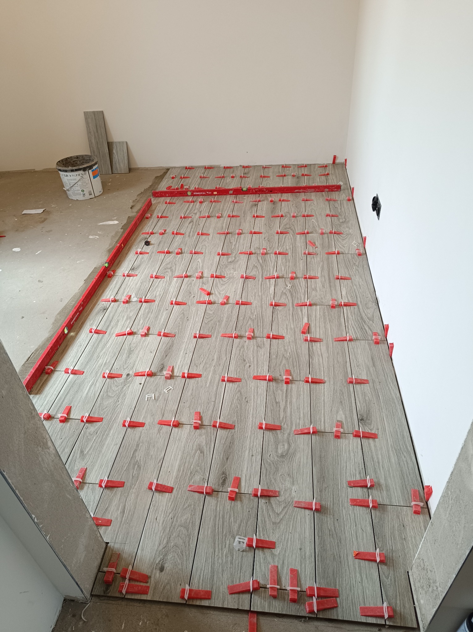 Ułożone płytki podłogowe imitujące drewno, z czerwonymi klipsami poziomującymi, poziomica przy ścianie, wiadro z farbą w tle. Wnętrze w trakcie wykańczania.