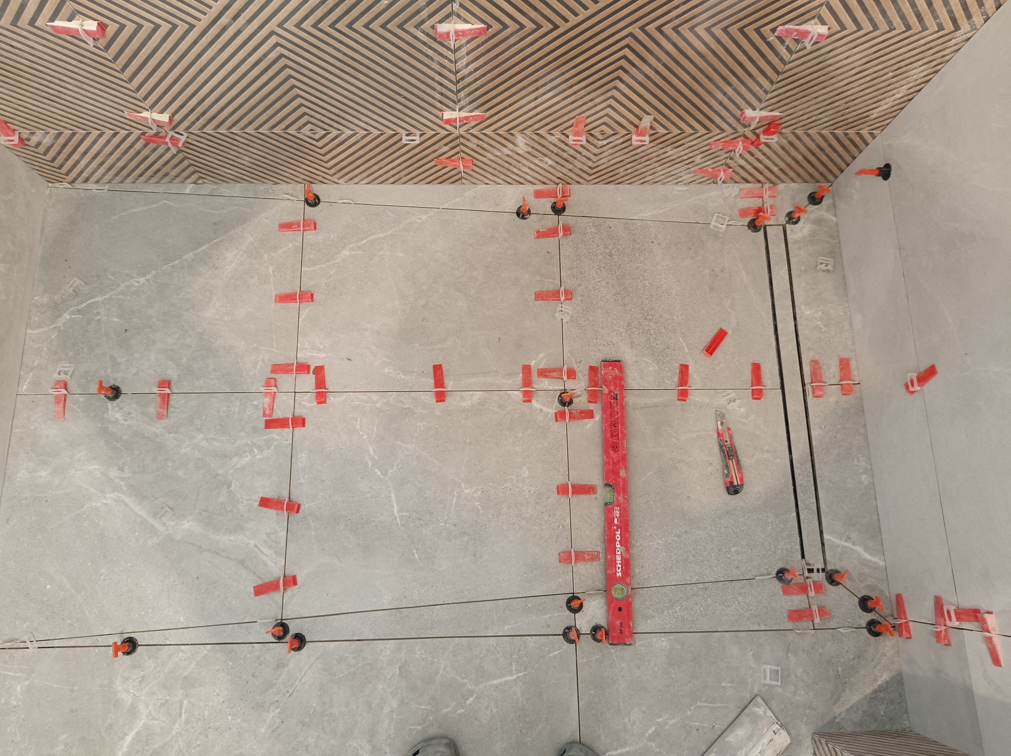 Układanie szarych płytek podłogowych i wzorzystych na suficie z użyciem poziomicy i klipsów. Widoczne fugi i narzędzia glazurnicze. Prace w toku.