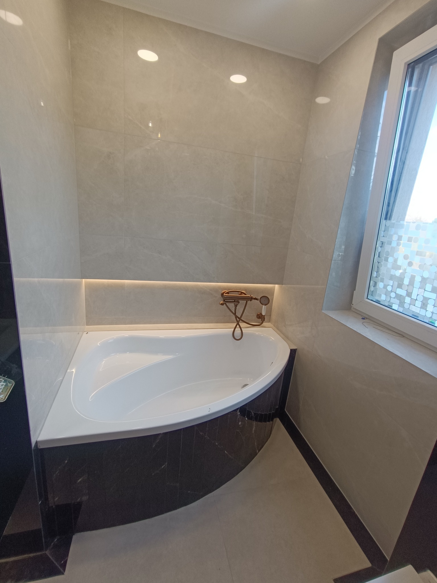 Wykończona łazienka z narożną wanną obudowaną ciemnym marmurem, jasne płytki na ścianach i podłodze, złota armatura, oświetlenie LED pod płytkami. Nowoczesny design.