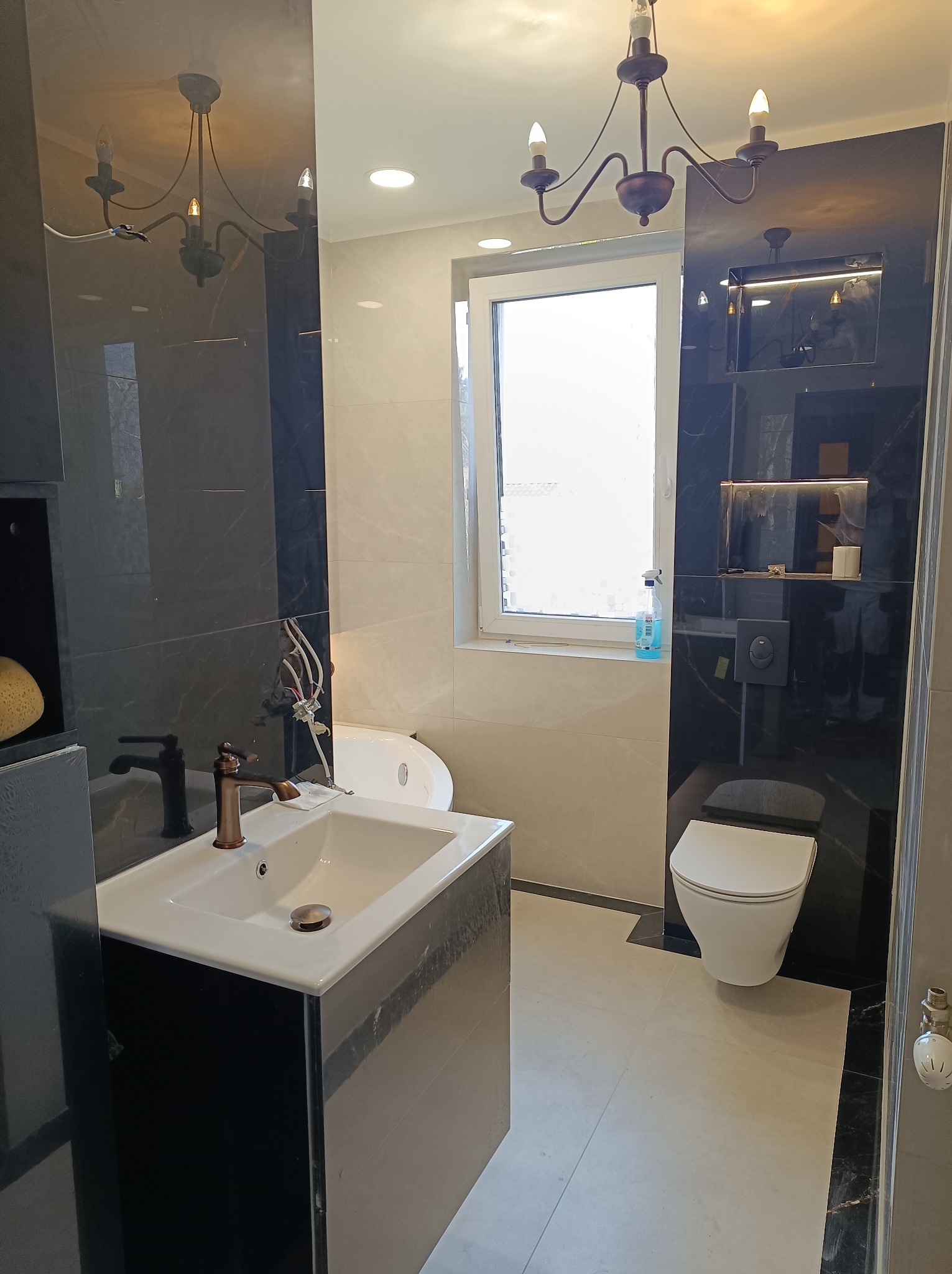 Elegancka łazienka po remoncie: biała umywalka z miedzianą armaturą, toaleta, wanna i czarne płytki imitujące marmur. Oświetlenie: żyrandol.