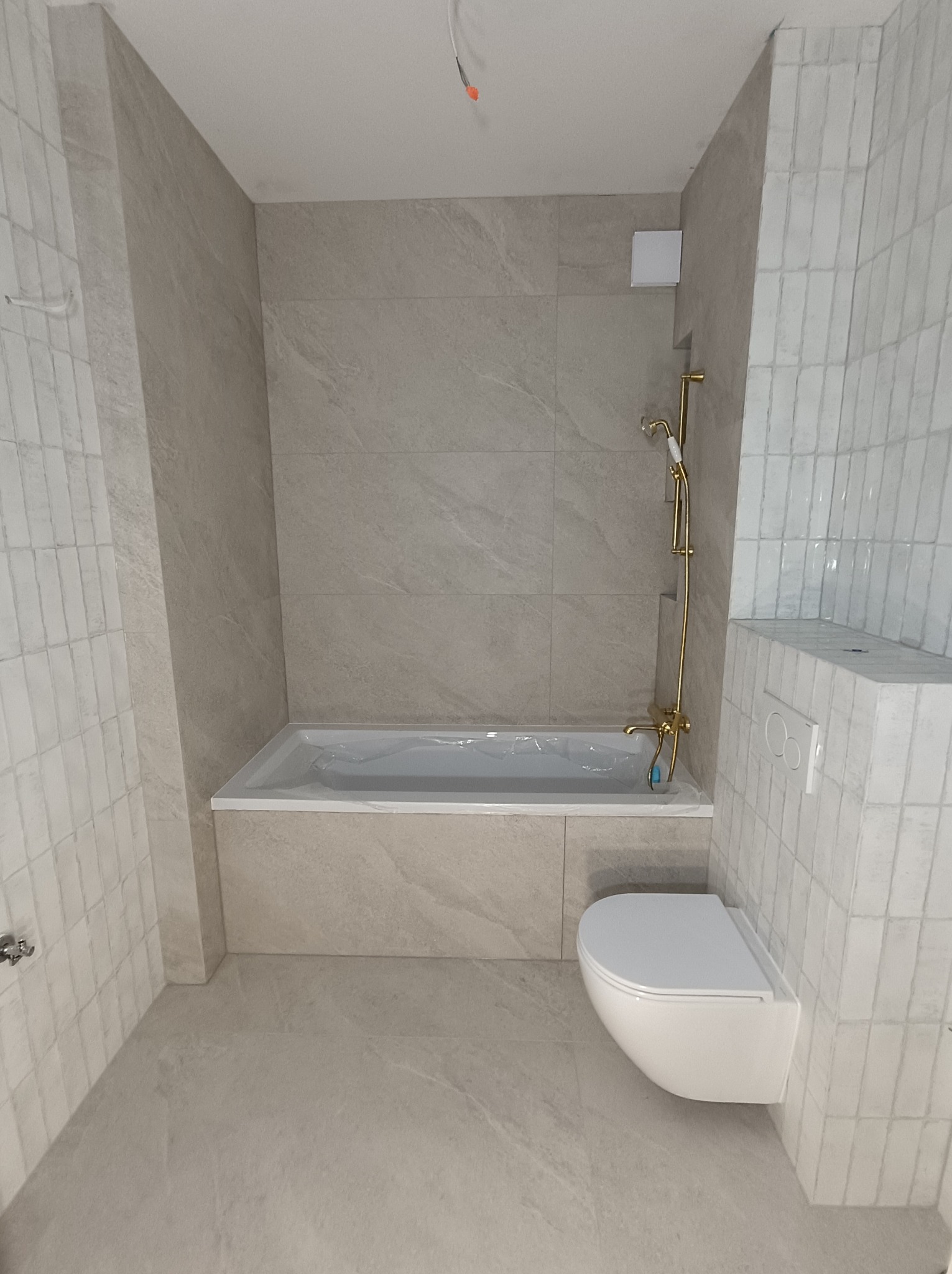 Jasna łazienka po remoncie: beżowe płytki imitujące kamień na ścianach i podłodze, biała wanna z obudową, toaleta podwieszana i złota armatura.