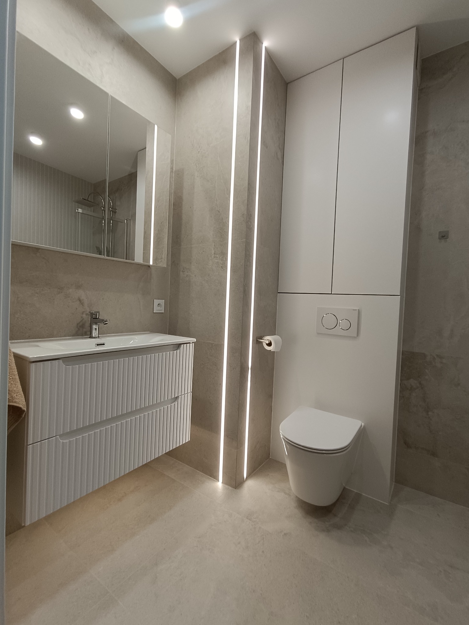 Nowoczesna łazienka z beżowymi płytkami, podświetlanymi pionowymi pasami LED, białą szafką z umywalką i podwieszaną toaletą. Minimalistyczny design.