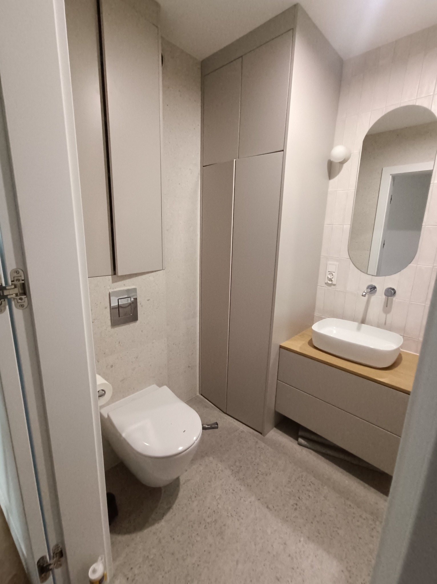 Nowoczesna łazienka w odcieniach szarości z wiszącą toaletą, umywalką nablatową i dużym lustrem owalnym. Minimalistyczny design, jasne płytki i szafki w zabudowie.