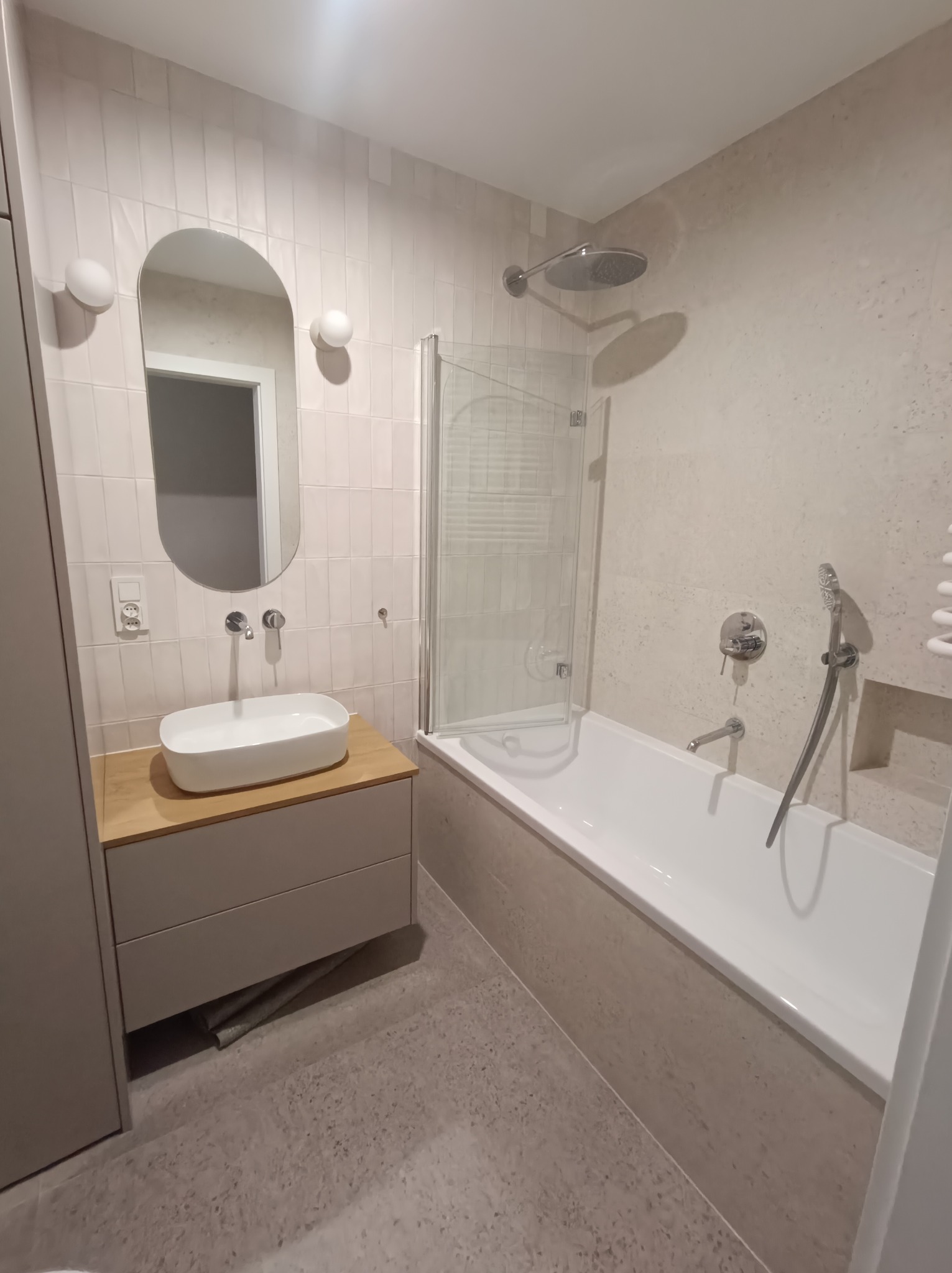 Jasna łazienka po remoncie: beżowe płytki na ścianach ułożone pionowo, owalne lustro nad umywalką, biała wanna z parawanem i szara podłoga z płytek.