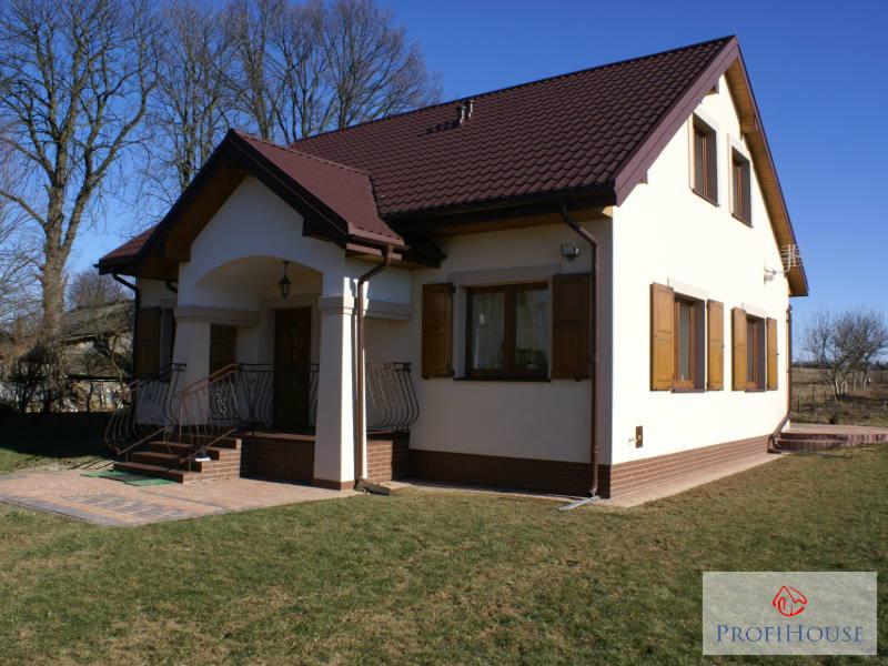 Elewacja domu jednorodzinnego z brązowym dachem i okiennicami, widok z ogrodu w słoneczny dzień. Dekoracyjne balustrady przy wejściu, cokół z cegły klinkierowej.