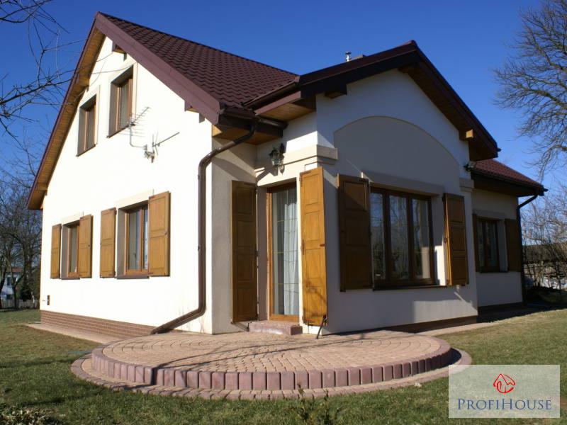 Jasny dom z brązowym dachem i okiennicami, widok z boku na okrągły taras z kostki. Błękitne niebo w tle, widać fragment ogrodu i drzewa.