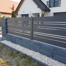 Nowoczesne, poziome przęsła ogrodzeniowe w kolorze grafitowym, zamontowane na podmurówce z szarej kostki łupanej, z białym grysem wzdłuż ogrodzenia i widokiem na dom z jasną elewacją w tle.
