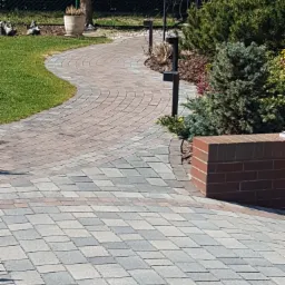 Zakrzywiony chodnik z kostki brukowej w dwóch odcieniach szarości i czerwieni, obok trawnik i rabata z roślinami iglastymi, zakończony niskim murkiem z cegły.