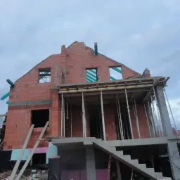 Surowy stan domu z czerwonej cegły, z widocznym szalunkiem i zbrojeniami. Budowa w toku, widać fundamenty i schody wejściowe.