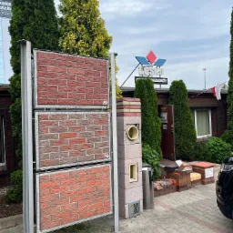 Ekspozycja trzech paneli imitujących mur z cegły w różnych odcieniach czerwieni, prezentowana przed budynkiem z szyldem 'LOSKO SPRZEDAŻY' i zielonymi tujami w tle.