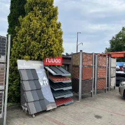 Ekspozycja różnych modeli pokryć dachowych i cegieł klinkierowych na zewnątrz, z logo producentów Wienerberger, Koramic i Ruukki, prezentowana na tle zielonych drzew i pochmurnego nieba.