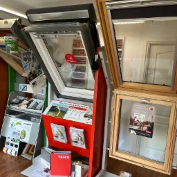 Ekspozycja okien dachowych w punkcie sprzedaży, prezentująca modele otwierane i z roletami, marki Fakro i Velux, wraz z materiałami informacyjnymi i cennikami.