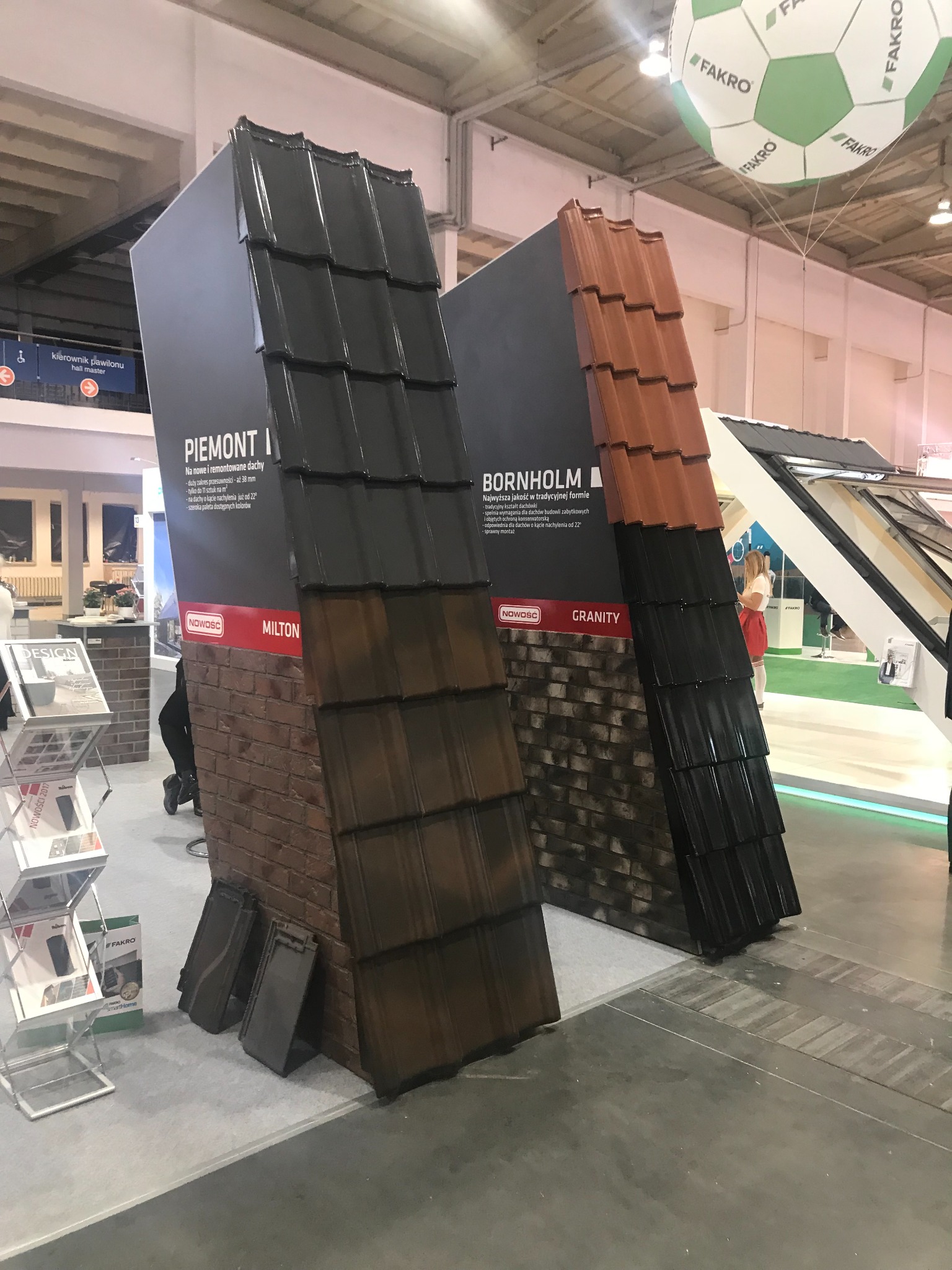 Ekspozycja dwóch modeli pokryć dachowych na stoisku targowym: ciemnobrązowy i czarny model 'Piemont I' z ceglaną ścianą w tle oraz pomarańczowo-czarny model 'Bornholm' z imitacją kamiennej ściany...