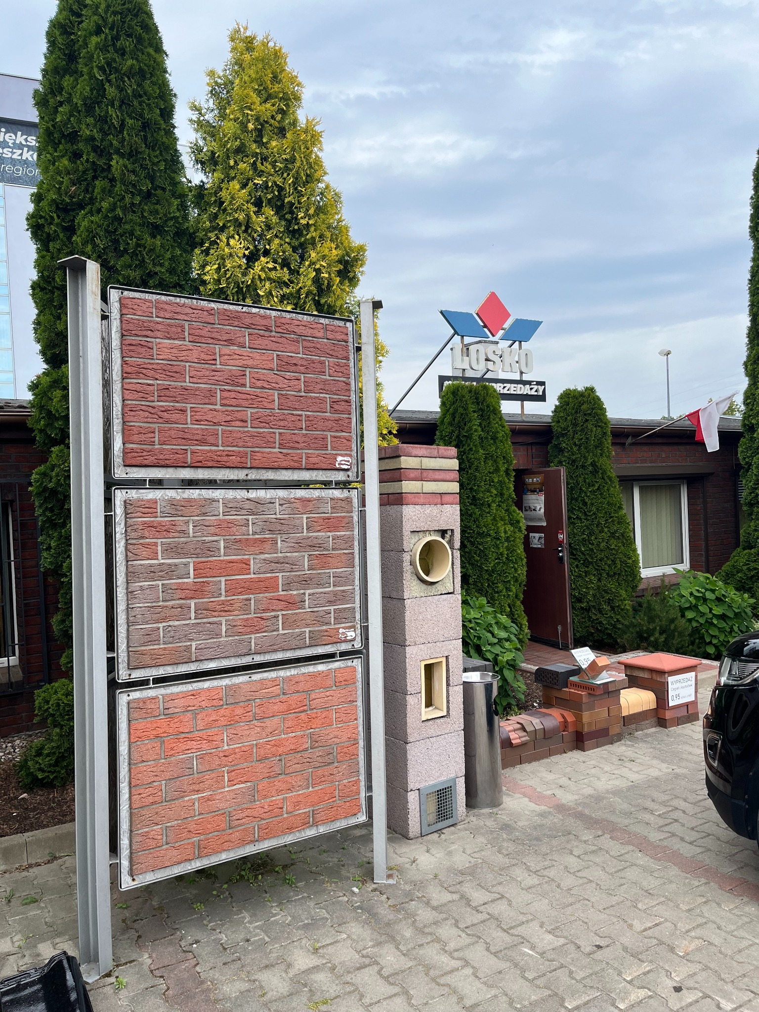 Ekspozycja trzech paneli imitujących mur z cegły w różnych odcieniach czerwieni, prezentowana przed budynkiem z szyldem 'LOSKO SPRZEDAŻY' i zielonymi tujami w tle.
