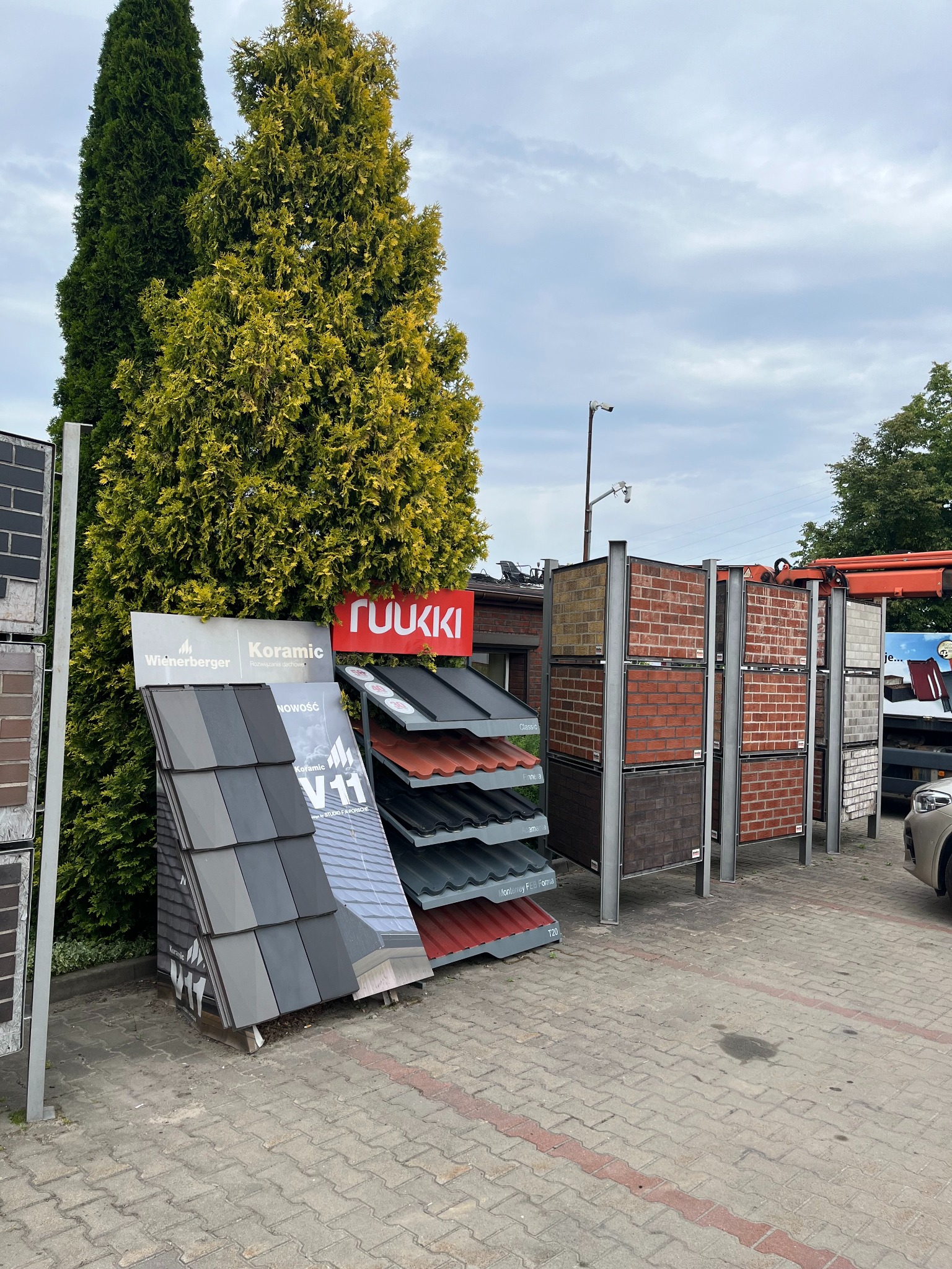 Ekspozycja różnych modeli pokryć dachowych i cegieł klinkierowych na zewnątrz, z logo producentów Wienerberger, Koramic i Ruukki, prezentowana na tle zielonych drzew i pochmurnego nieba.