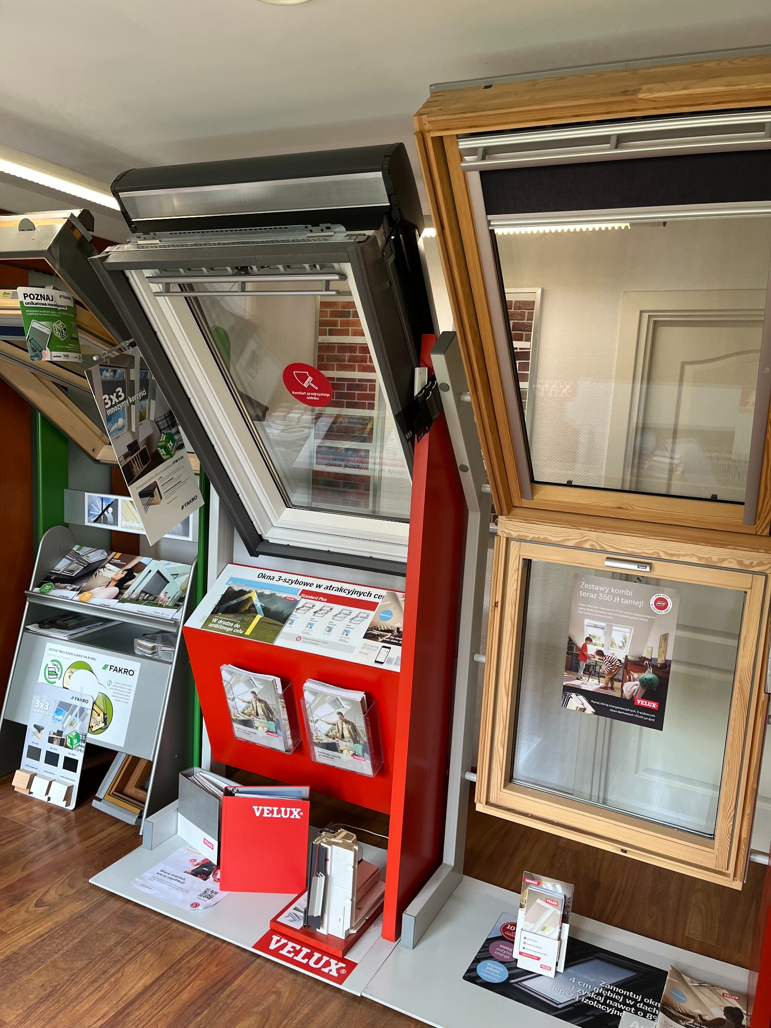 Ekspozycja okien dachowych w punkcie sprzedaży, prezentująca modele otwierane i z roletami, marki Fakro i Velux, wraz z materiałami informacyjnymi i cennikami.