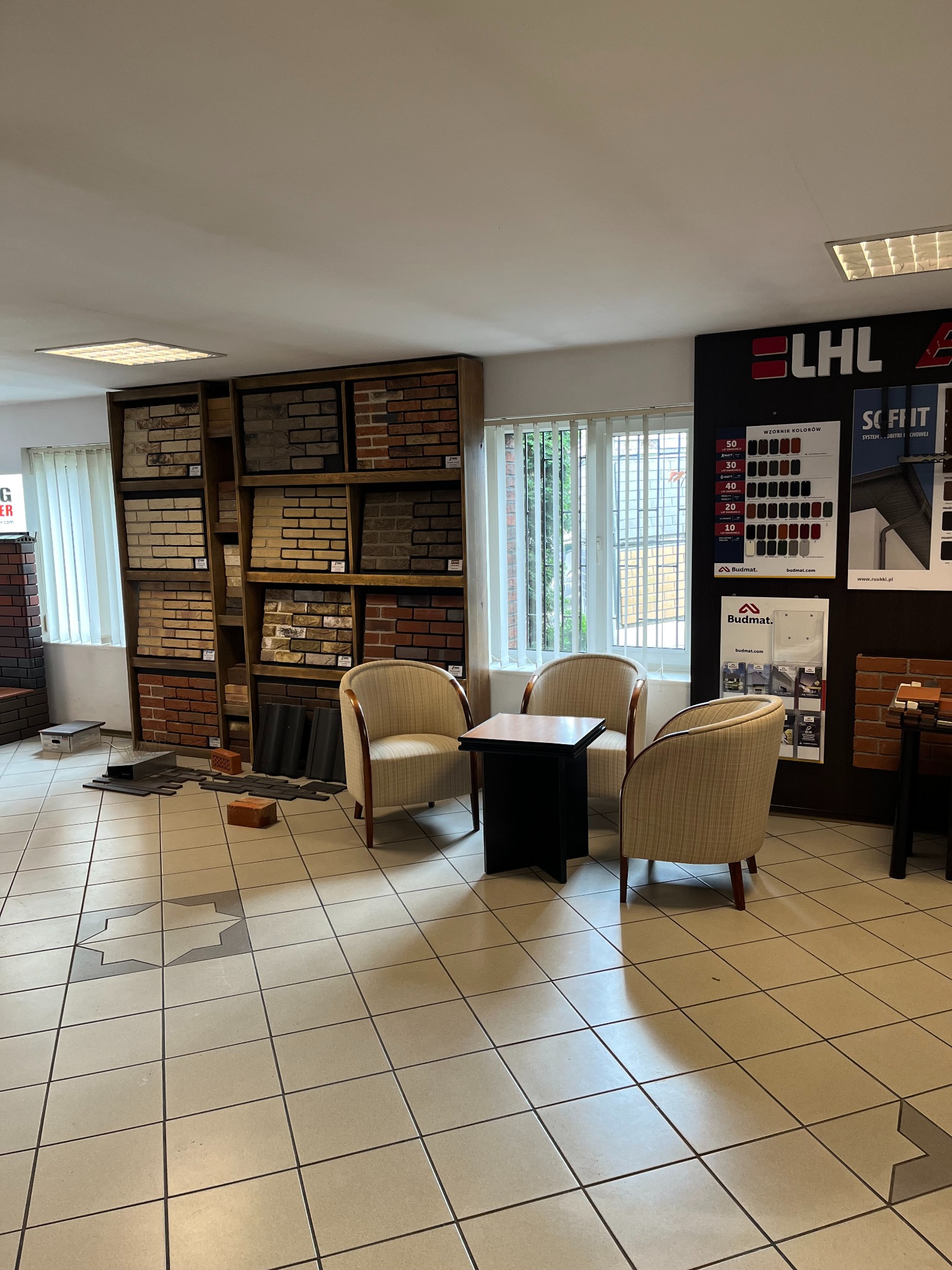 Ekspozycja różnorodnych materiałów budowlanych w sklepie, w tym cegieł i dachówek, prezentowana na półkach w tle, z trzema fotelami i małym stolikiem na pierwszym planie.