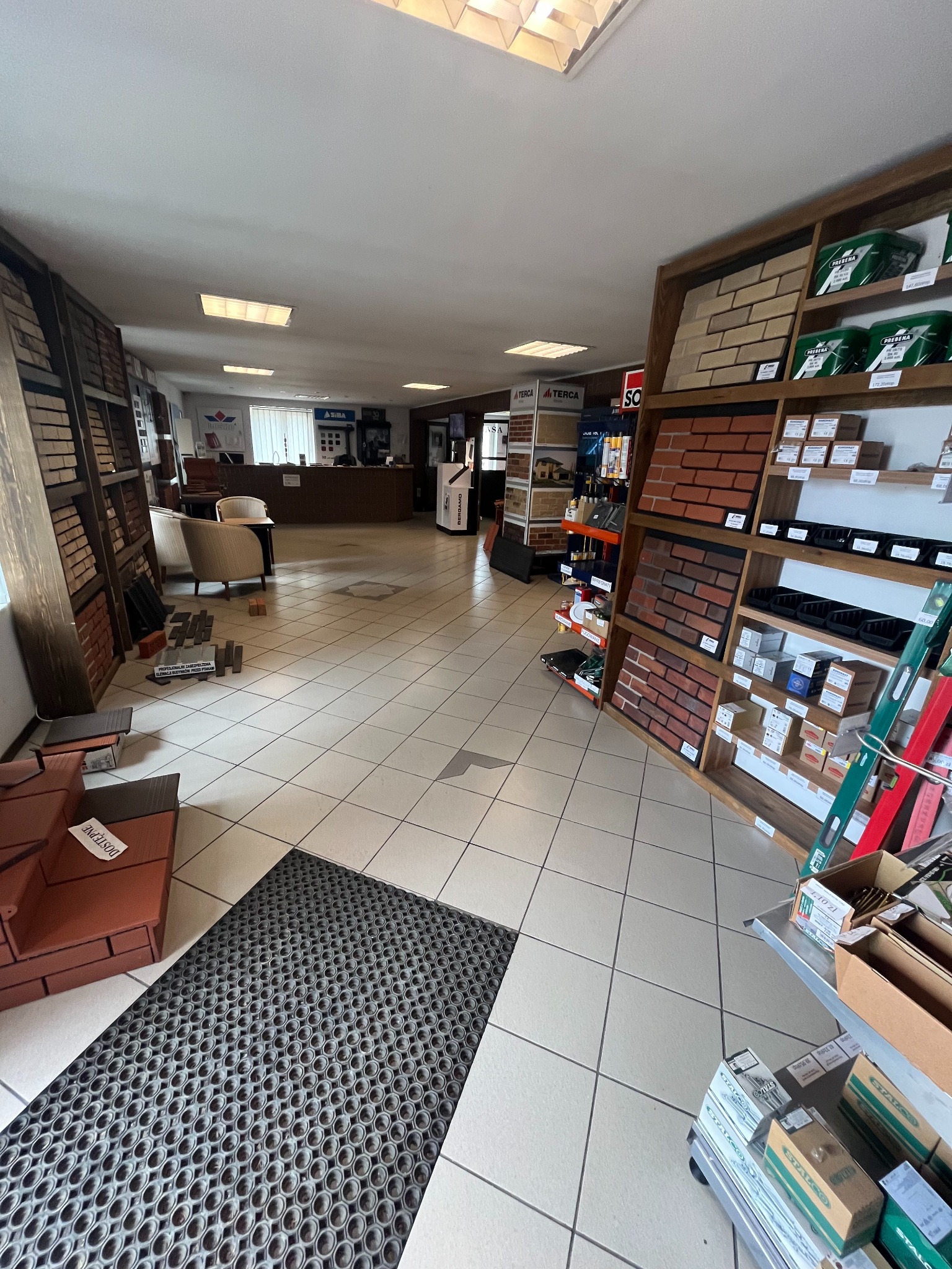 Wnętrze sklepu z materiałami budowlanymi, prezentujące ekspozycję różnorodnych cegieł i materiałów wykończeniowych na drewnianych regałach, widoczne również materiały do mocowania i narzędzia.