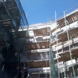 Pracownik w kasku i kamizelce na rusztowaniu przy białej ścianie budynku w trakcie prac budowlanych. Widok z dołu, błękitne niebo.