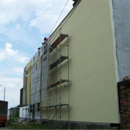 DUMAjd - Ocieplanie elewacji budynku wielorodzinnego z użyciem rusztowania, widoczny fragment nowej, jasnej elewacji oraz stara, surowa ściana. Na rusztowaniu pracuje osoba.