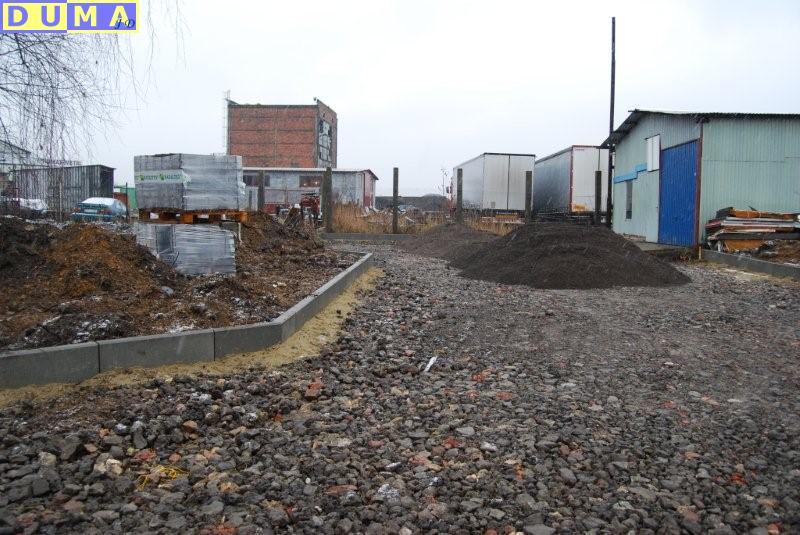 Układanie krawężników i przygotowanie podłoża z gruzu pod brukowaną ścieżkę na terenie przemysłowym, widoczne palety z kostką brukową, hałdy ziemi i budynki gospodarcze w tle.
