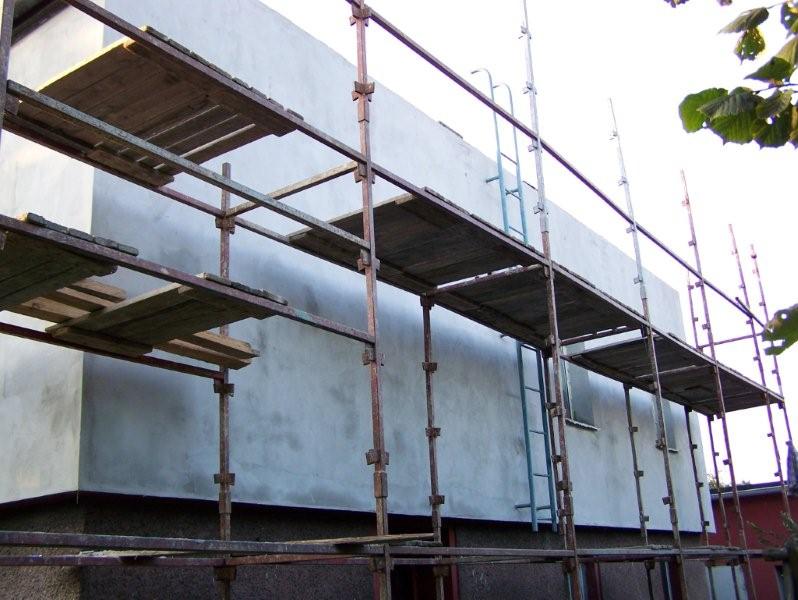 Budynek w trakcie ocieplania, widoczne rusztowania i fragment elewacji pokrytej szarym tynkiem.