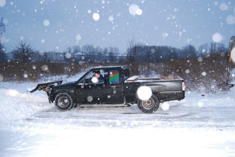 Czarny pickup z zamontowanym pługiem odśnieżającym, odśnieżanie drogi podczas obfitych opadów śniegu, widoczny kierowca w kabinie.