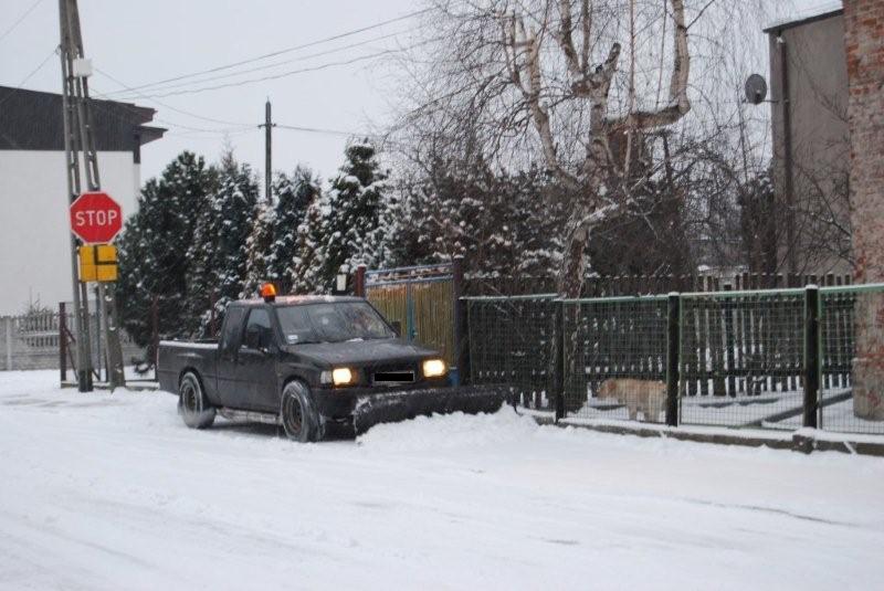 Czarny pickup z pługiem odśnieżającym na przodzie, usuwający śnieg z drogi w zimowy dzień, z widocznym znakiem stop i ogrodzeniem w tle.