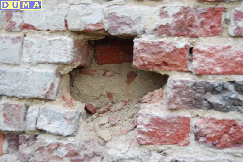 Uszkodzona, stara ściana z czerwonej cegły z ubytkiem wypełnionym zaprawą, widoczne fragmenty cegieł wewnątrz ubytku.