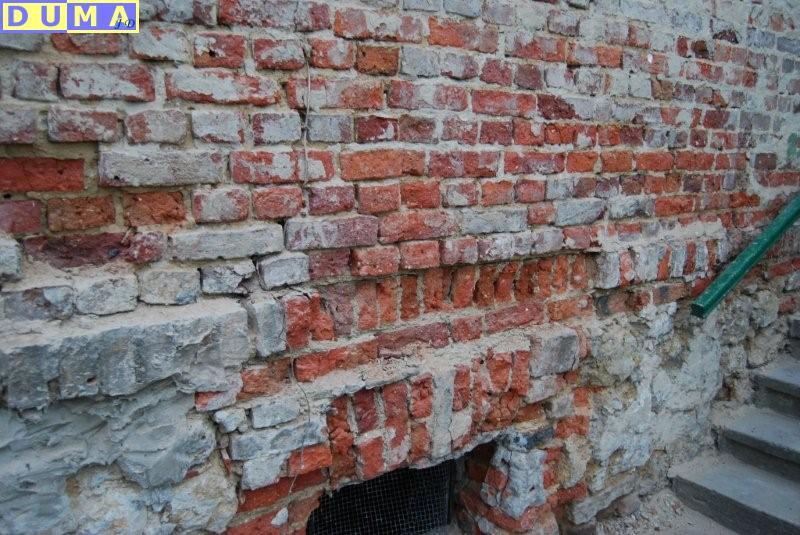 Fragment zniszczonej, starej ściany z czerwonej cegły z ubytkami i naprawami, widoczna siatka zabezpieczająca w otworze oraz fragment schodów z zieloną barierką.