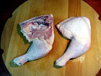 Dwie surowe nogi kurczaka na okrągłej drewnianej desce do krojenia. Jedna noga przekrojona, ukazująca strukturę mięsa i kości.
