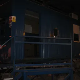 Zewnętrzny widok niebieskiego kontenera biurowego z metalowymi schodami i poręczami, widoczne znaki ostrzegawcze na ścianie i dziura w poszyciu, słabe oświetlenie.