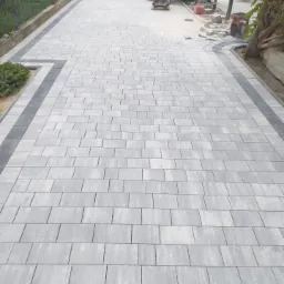 Układanie szarej kostki brukowej prostokątnej z ciemnoszarą obwódką, w tle żółty samochód dostawczy i ubijarka wibracyjna.