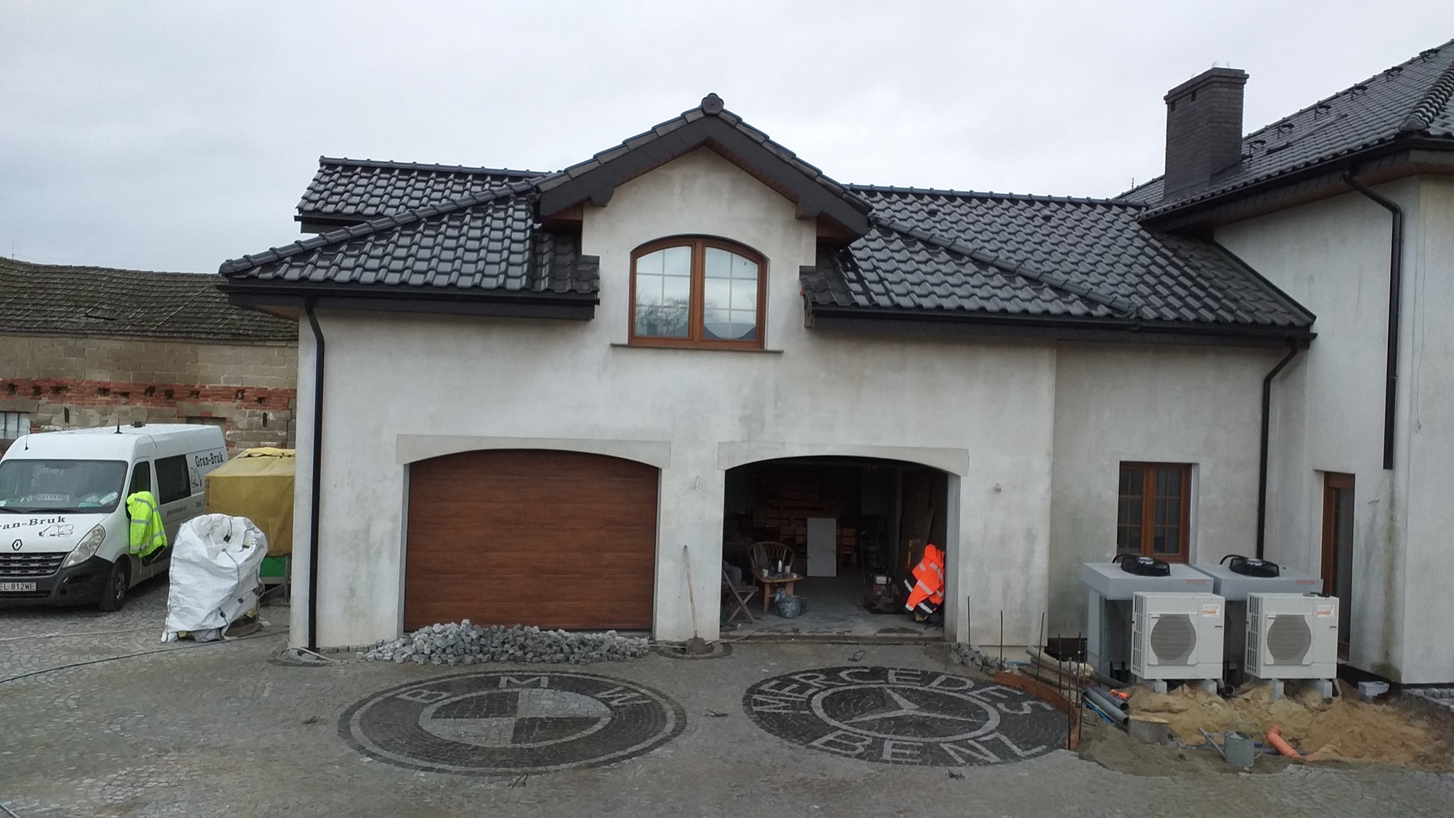 Dwustanowiskowy garaż domu jednorodzinnego z mozaiką z kostki brukowej przedstawiającą logo BMW i Mercedes-Benz na podjeździe, obok zaparkowany bus firmy brukarskiej.