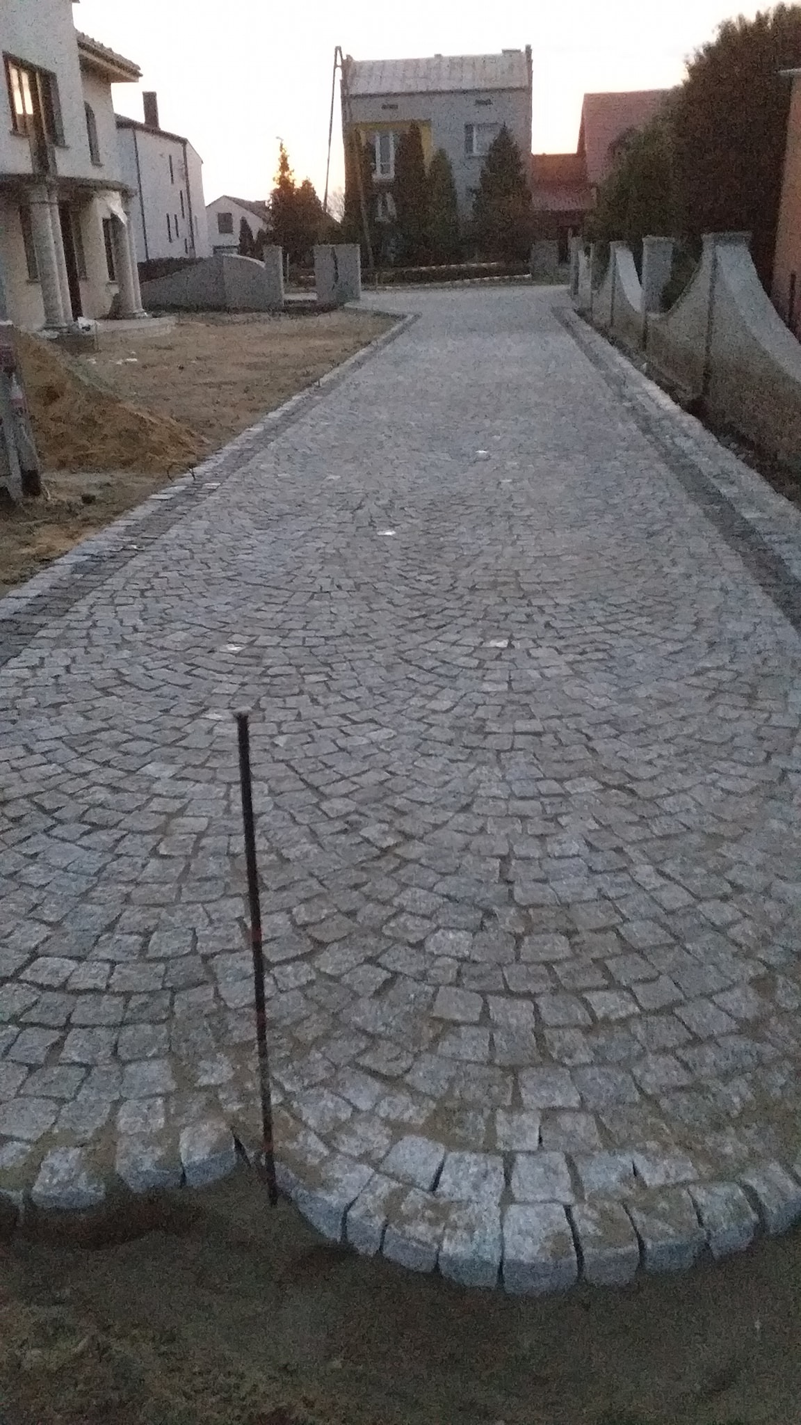Ułożona droga z szarej kostki granitowej w kształcie wachlarza, widoczne budynki w tle i metalowy pręt wbity w ziemię.